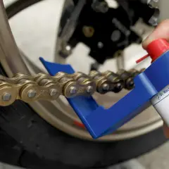 Hand sprüht mit einer Dose ein Motorrad Kette, um sie zu fetten. Ein blauer Kettenreiniger hilft dabei.