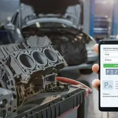 Hand hält Smartphone mit Fehlercode P0266 für einen Mercedes, daneben ein zerlegter Motorblock in einer Werkstatt.