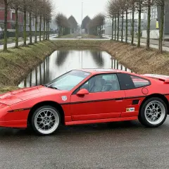 Roter Pontiac Fiero GT mit Felgen steht auf nasser Straße neben einem Kanal.