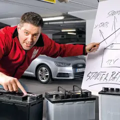 Mann erklärt den Wechsel einer Audi Q5 Batterie. Er zeigt auf eine Zeichnung einer Starterbatterie und auf drei Batterien.