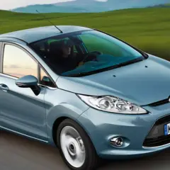 Blauer Ford Fiesta fährt auf einer Straße mit grünen Hügeln im Hintergrund.