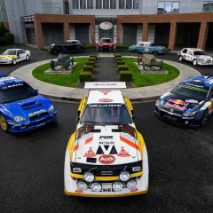 Blauer Subaru WRC Rallye-Bolide neben einem Audi Quattro und einem VW Polo. Historische Rennwagen im Hintergrund.