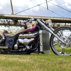 Ein glänzender Boss Hoss V8-Chopper mit lila-goldener Lackierung und einem riesigen V8-Motor steht auf einer Wiese vor einem Flugzeugwrack.