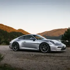 Silberner Porsche 911 ST parkt am Ufer eines Sees, umgeben von bewaldeten Hügeln im warmen Abendlicht.