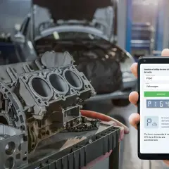 Ein Smartphone zeigt den Fehlercode P1641 für einen Volkswagen an, während im Hintergrund ein zerlegter Motor liegt.