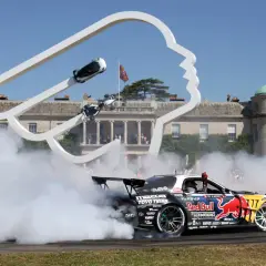 Drift-Auto mit Red Bull-Logo erzeugt Rauchwolke vor einer Skulptur und historischem Gebäude. Ein Fahrer winkt, während er den Goodwood Record bricht.
