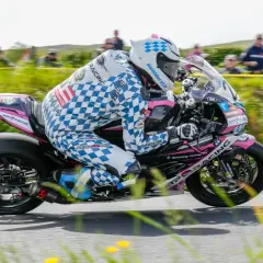 Motorradrennfahrer in kariertem Anzug fährt auf einer Rennstrecke, Zuschauer im Hintergrund.