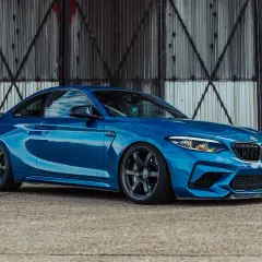 Ein blauer BMW F87 M2 mit dunklen Felgen steht vor einer rostigen Metallwand.