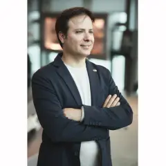 Christopher Pagani, mit verschränkten Armen und einem Lächeln, blickt nach rechts. Er trägt einen dunkelblauen Blazer über einem weißen T-Shirt.