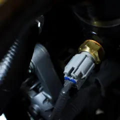 Ein p0118 vw Sensor mit grauem Stecker und Messinggewinde, eingebettet in dunkle Motorraumkabel.