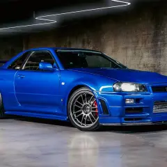 Ein blauer Nissan Skyline R34 GT-R mit Spoiler und auffälligen Felgen steht in einer Garage.