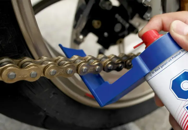 Hand sprüht mit einer Dose ein Motorrad Kette, um sie zu fetten. Ein blauer Kettenreiniger hilft dabei.