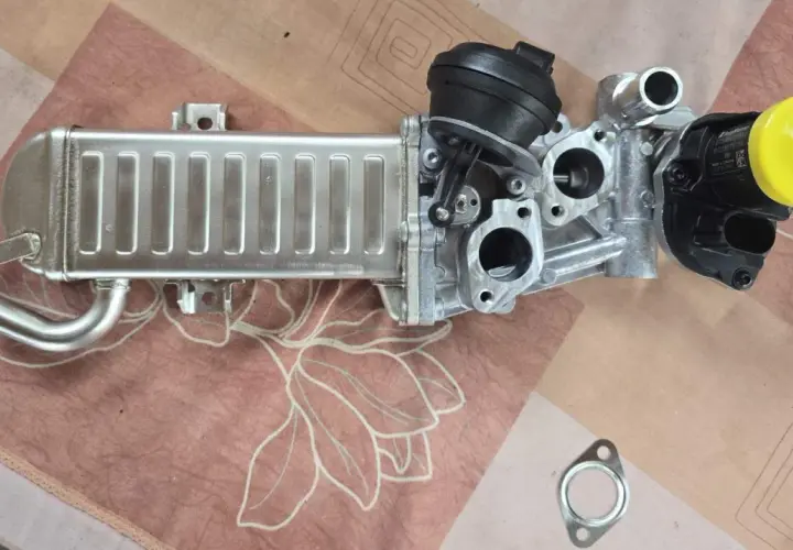 VW AGR-Kühler mit schwarzem Stellmotor und gelber Kappe auf gemustertem Untergrund.