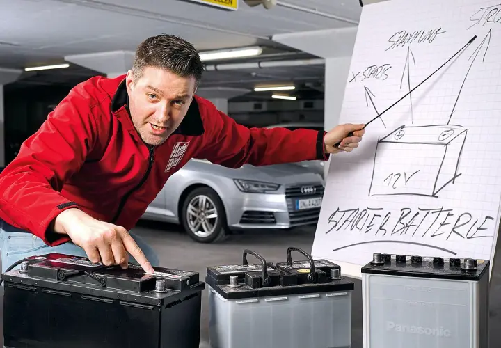Mann erklärt den Wechsel einer Audi Q5 Batterie. Er zeigt auf eine Zeichnung einer Starterbatterie und auf drei Batterien.