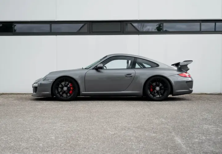 Grauer Porsche 997 GT3 RS mit schwarzem Flügel und roten Bremssätteln steht vor einer weißen Wand.
