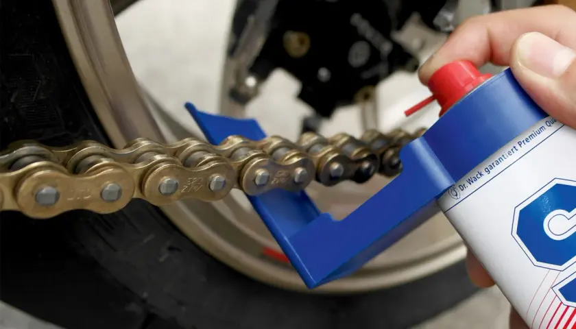 Hand sprüht mit einer Dose ein Motorrad Kette, um sie zu fetten. Ein blauer Kettenreiniger hilft dabei.