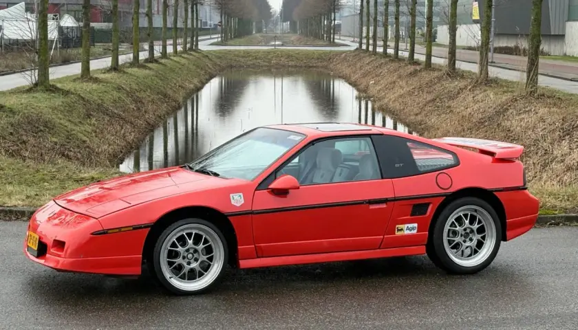 Roter Pontiac Fiero GT mit Felgen steht auf nasser Straße neben einem Kanal.