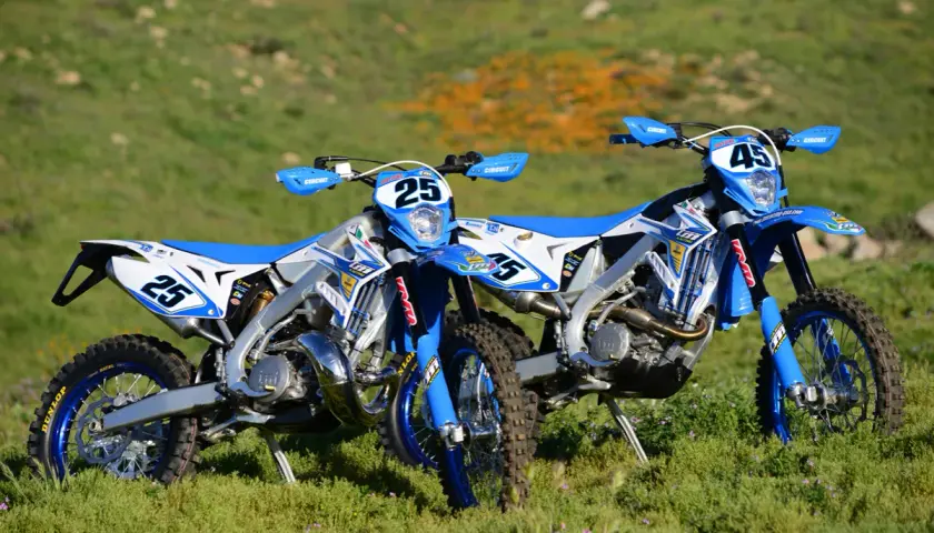 Zwei blaue Enduro-Motorräder, ein 2-Takter und ein 4-Takter, stehen auf einer grünen Wiese.