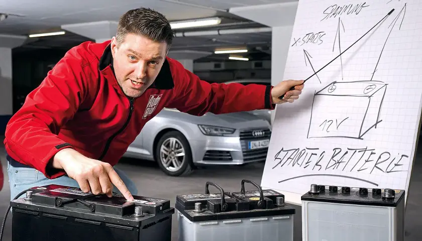 Mann erklärt den Wechsel einer Audi Q5 Batterie. Er zeigt auf eine Zeichnung einer Starterbatterie und auf drei Batterien.