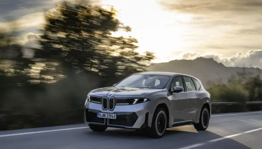 Silberner BMW iX3 fährt auf einer Straße im Sonnenuntergang. Informieren Sie sich über die Lieferzeiten des BMW iX3.