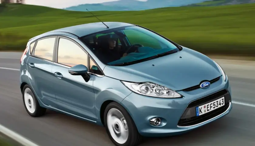 Blauer Ford Fiesta fährt auf einer Straße mit grünen Hügeln im Hintergrund.