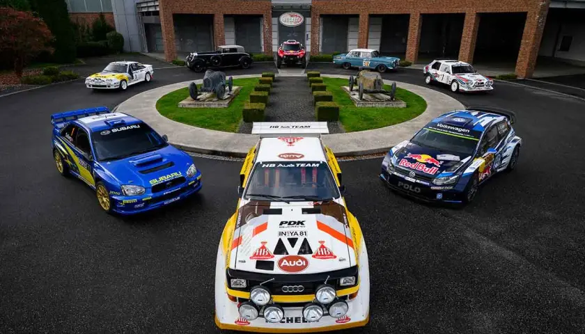 Blauer Subaru WRC Rallye-Bolide neben einem Audi Quattro und einem VW Polo. Historische Rennwagen im Hintergrund.
