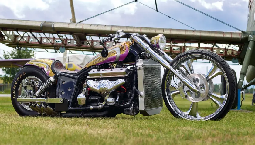 Ein glänzender Boss Hoss V8-Chopper mit lila-goldener Lackierung und einem riesigen V8-Motor steht auf einer Wiese vor einem Flugzeugwrack.