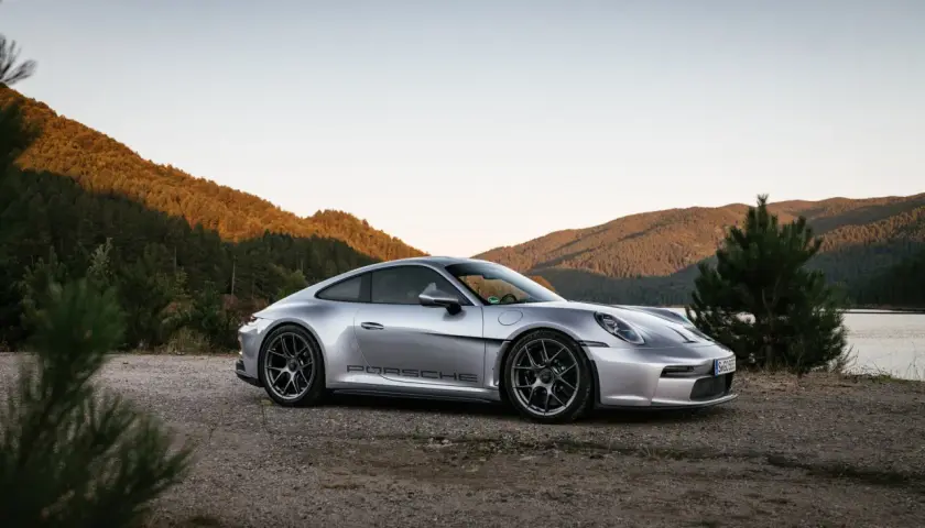 Silberner Porsche 911 ST parkt am Ufer eines Sees, umgeben von bewaldeten Hügeln im warmen Abendlicht.
