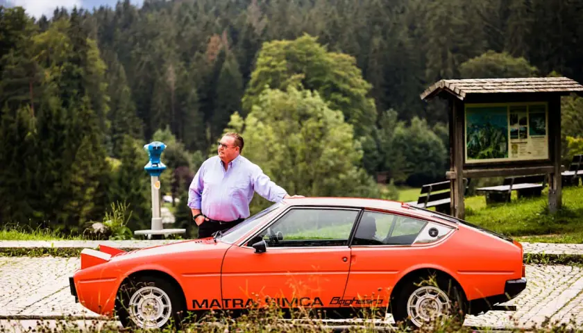 Ein Mann lehnt an seinem leuchtend orangen Simca Matra Bagheera. Im Hintergrund ein dichter Nadelwald.