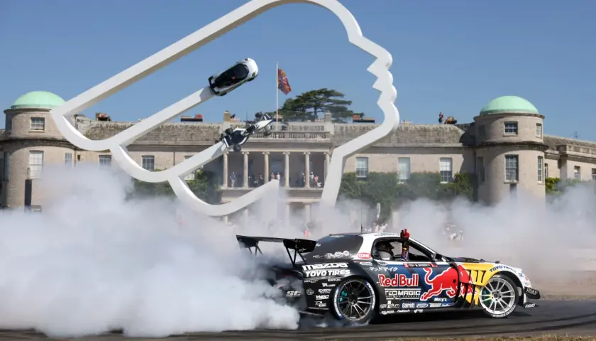 Drift-Auto mit Red Bull-Logo erzeugt Rauchwolke vor einer Skulptur und historischem Gebäude. Ein Fahrer winkt, während er den Goodwood Record bricht.