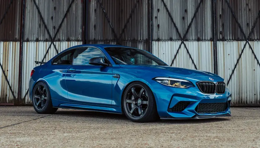 Ein blauer BMW F87 M2 mit dunklen Felgen steht vor einer rostigen Metallwand.