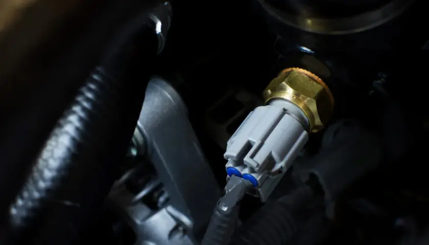 Ein p0118 vw Sensor mit grauem Stecker und Messinggewinde, eingebettet in dunkle Motorraumkabel.