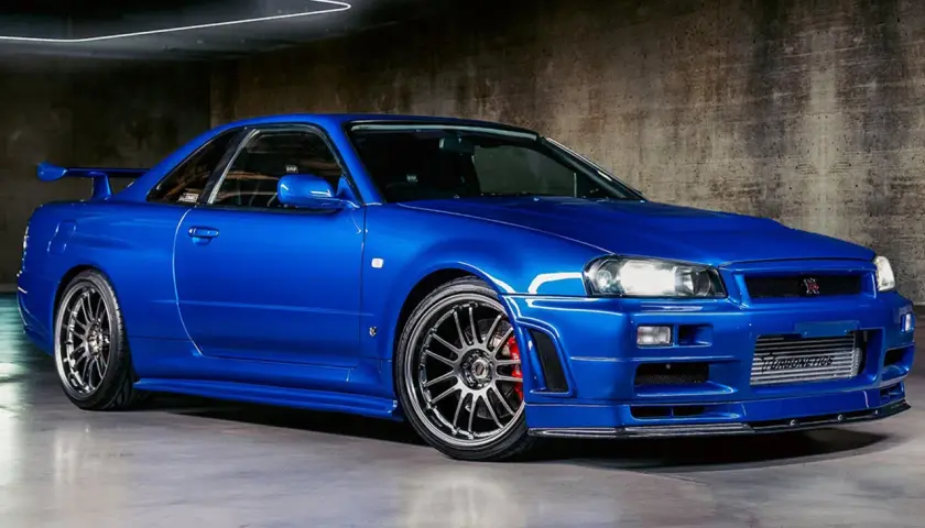 Ein blauer Nissan Skyline R34 GT-R mit Spoiler und auffälligen Felgen steht in einer Garage.