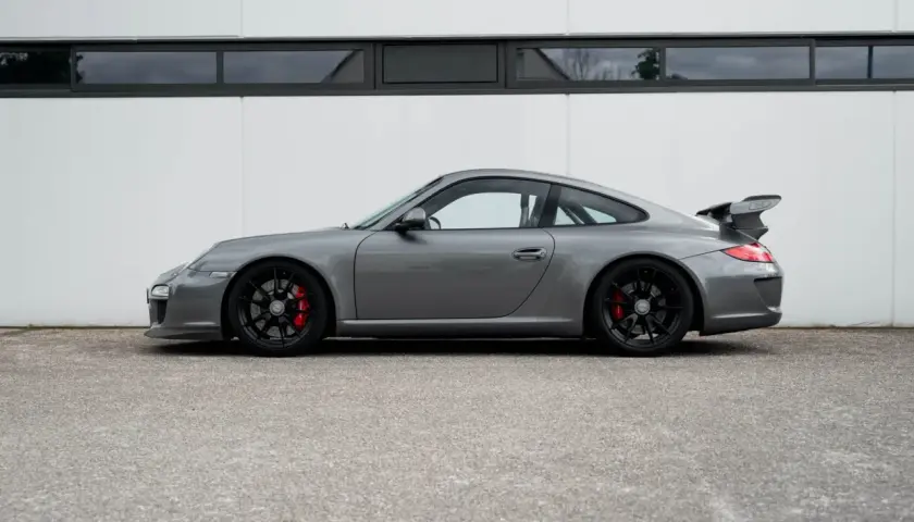 Grauer Porsche 997 GT3 RS mit schwarzem Flügel und roten Bremssätteln steht vor einer weißen Wand.