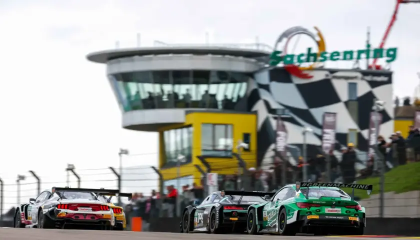 Rennwagen rasen auf dem Sachsenring. Ein grüner BMW mit #GREENMACHINE-Schriftzug und ein roter Ford Mustang liefern sich ein spannendes DTM-Rennen.