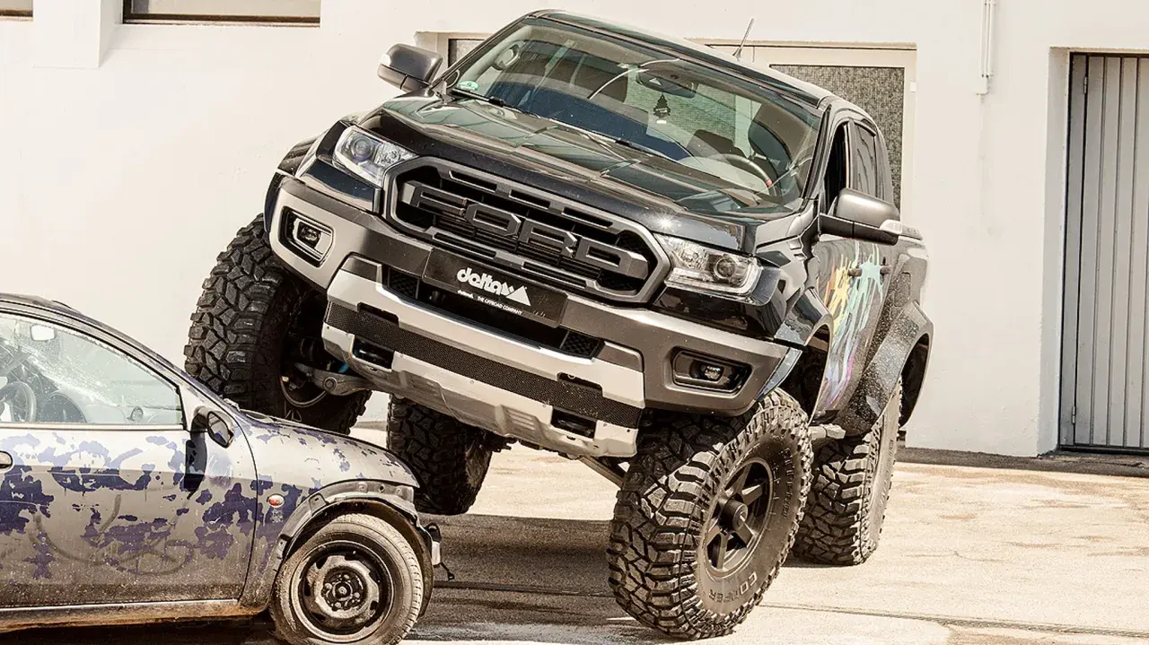 Schwarzer Ford Ranger mit **Ford Duratorq**-Motor überfährt ein kleines Auto. Offroad-Umbau mit riesigen Reifen.