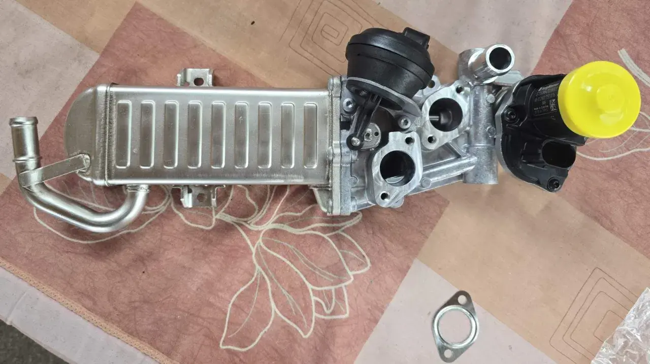 VW AGR-Kühler mit schwarzem Stellmotor und gelber Kappe auf gemustertem Untergrund.