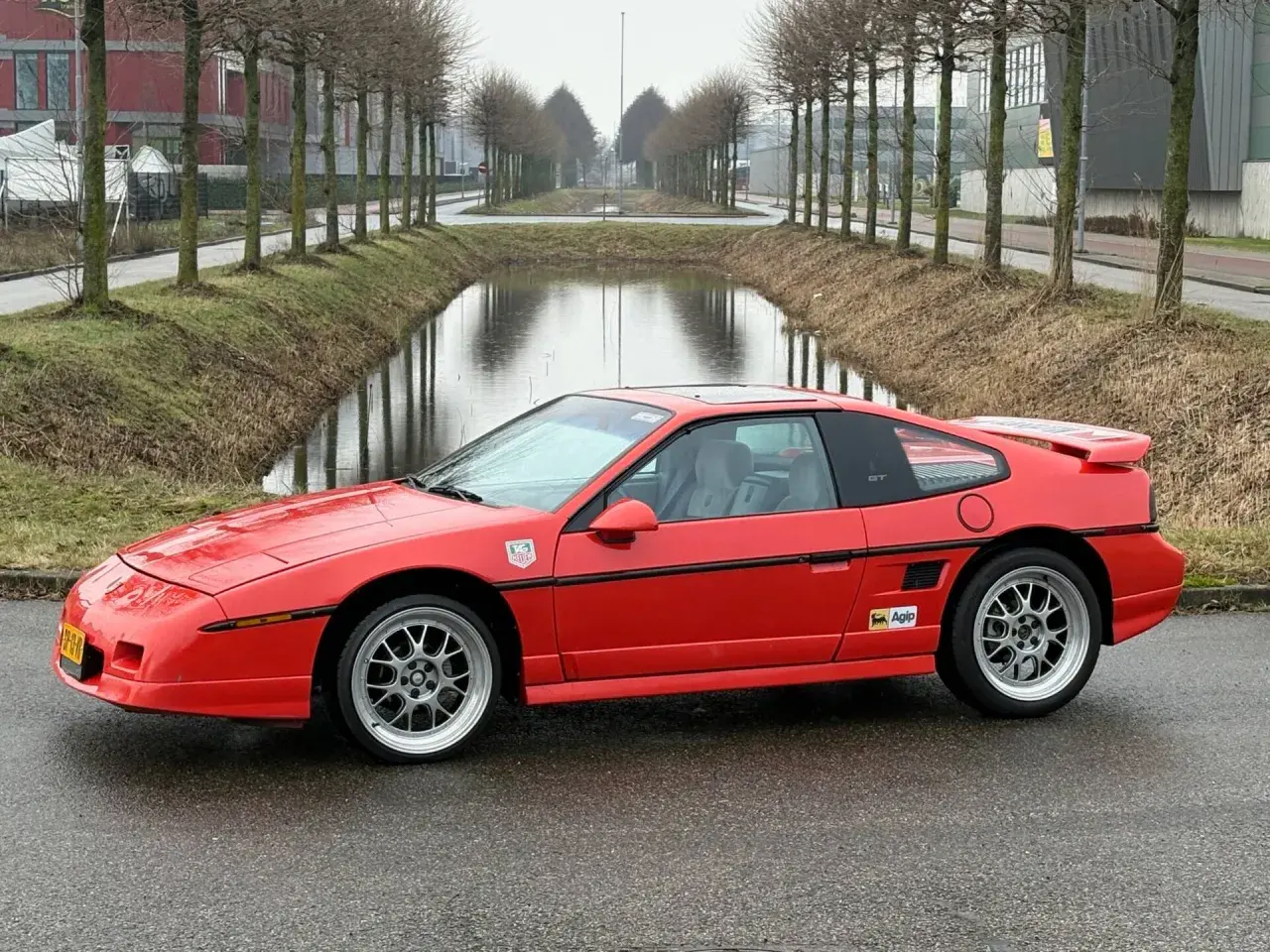 Roter Pontiac Fiero GT mit Felgen steht auf nasser Straße neben einem Kanal.