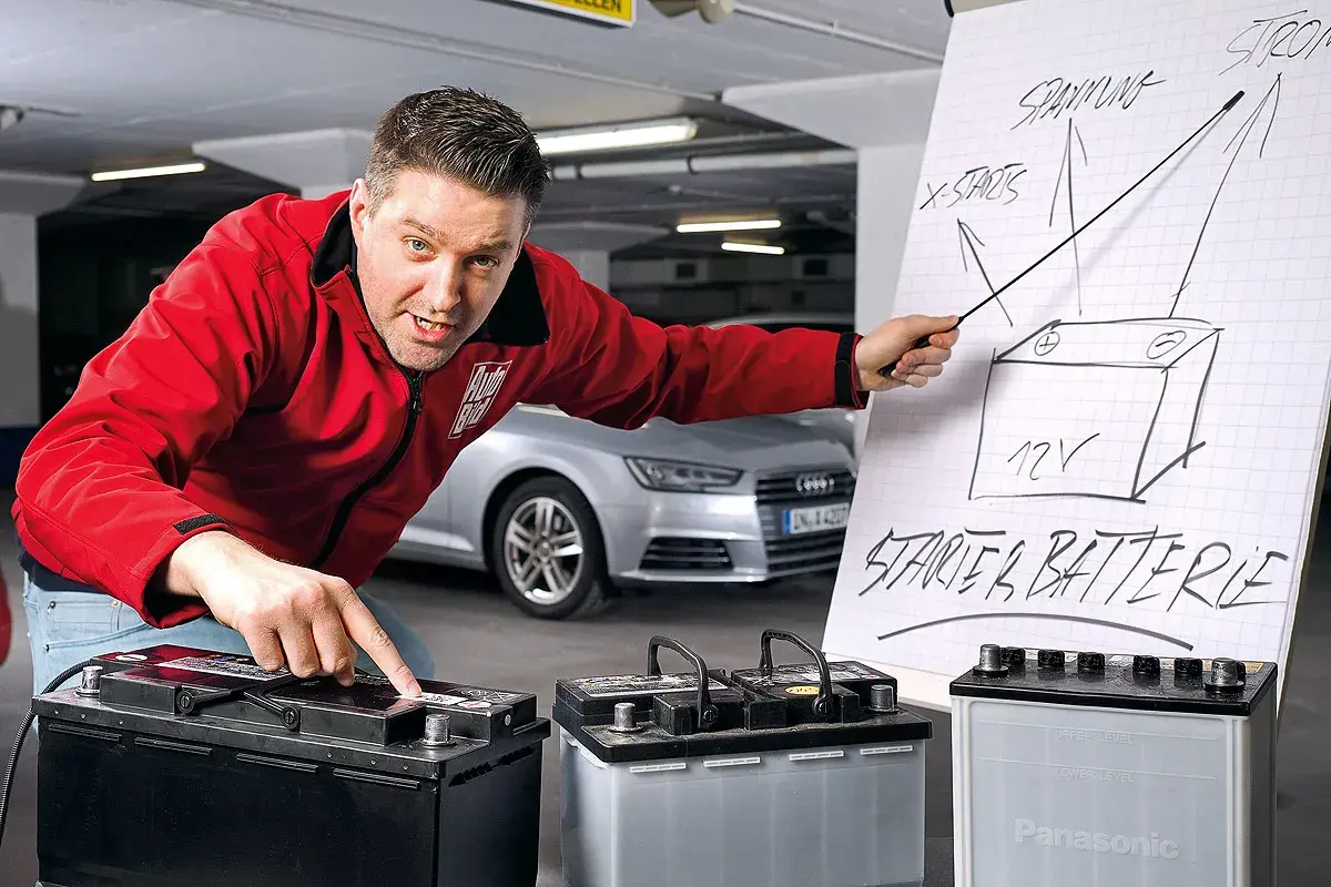 Mann erklärt den Wechsel einer Audi Q5 Batterie. Er zeigt auf eine Zeichnung einer Starterbatterie und auf drei Batterien.