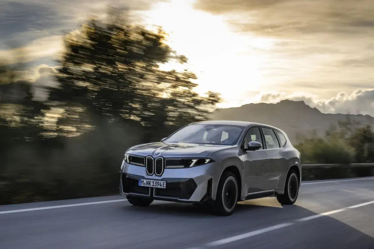 Silberner BMW iX3 fährt auf einer Straße im Sonnenuntergang. Informieren Sie sich über die Lieferzeiten des BMW iX3.