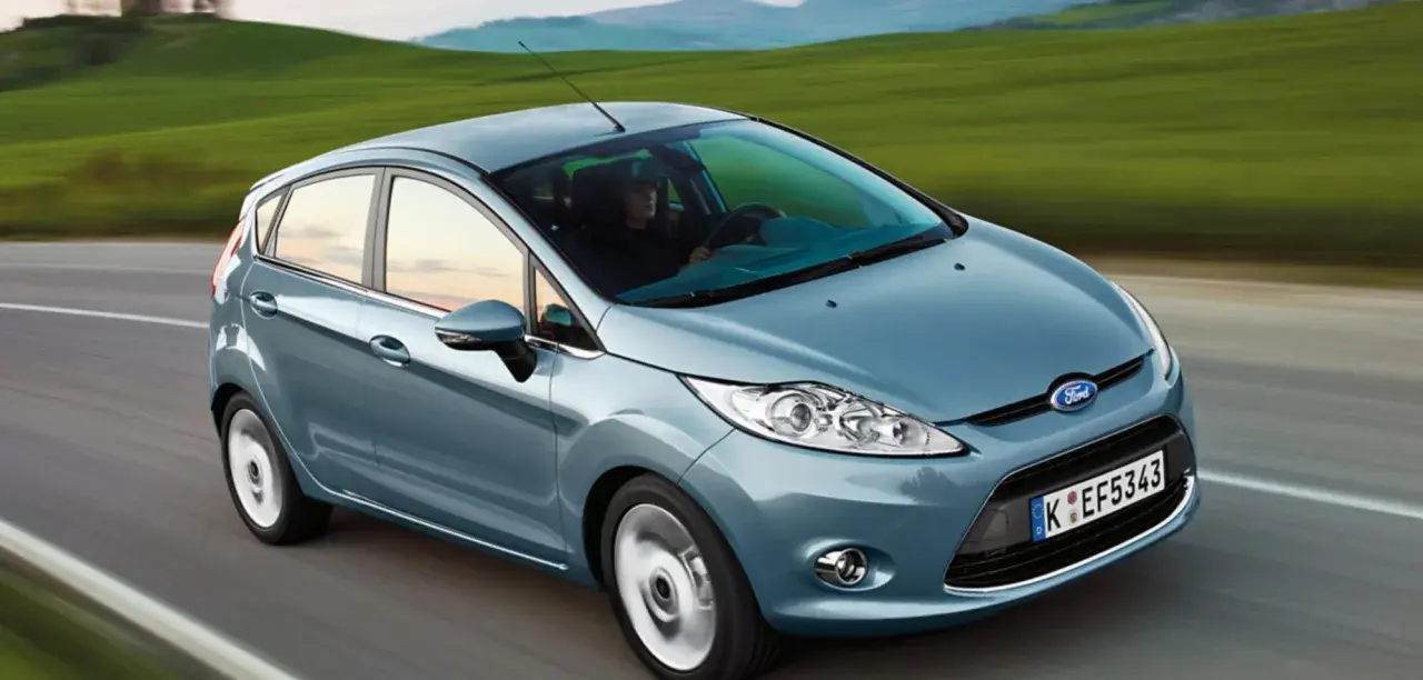 Blauer Ford Fiesta fährt auf einer Straße mit grünen Hügeln im Hintergrund.