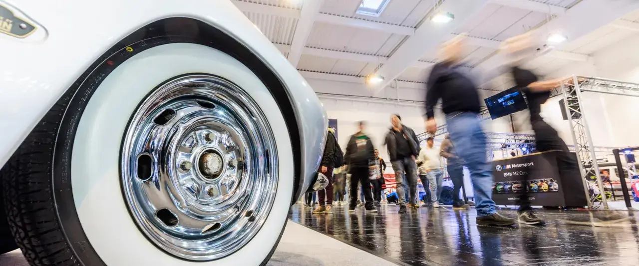 Detailaufnahme eines Oldtimer-Reifens mit Chromfelge auf einer motorsport expo. Besucher bewegen sich im Hintergrund.