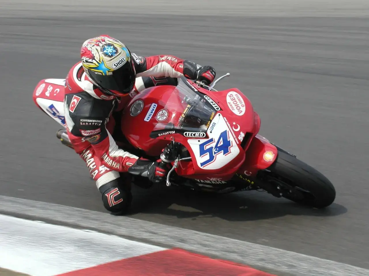 Rennfahrer Kenan Sofuoğlu in roter Lederkombi auf roter Yamaha mit Startnummer 54, in Schräglage auf der Rennstrecke.