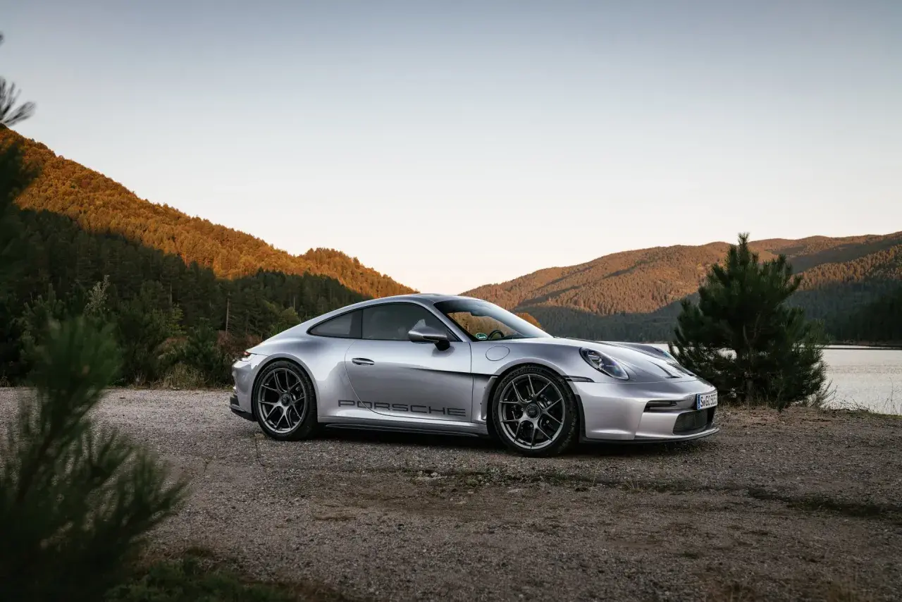 Silberner Porsche 911 ST parkt am Ufer eines Sees, umgeben von bewaldeten Hügeln im warmen Abendlicht.