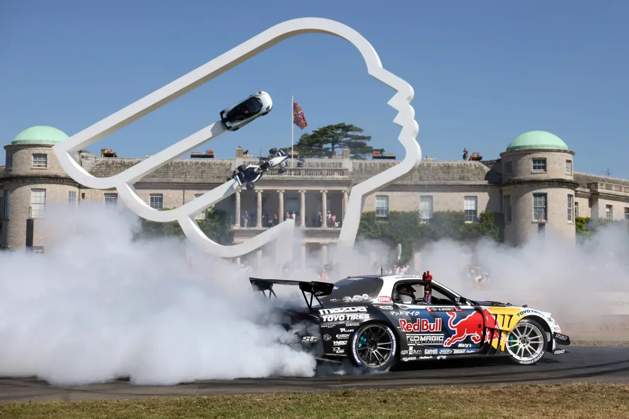 Drift-Auto mit Red Bull-Logo erzeugt Rauchwolke vor einer Skulptur und historischem Gebäude. Ein Fahrer winkt, während er den Goodwood Record bricht.