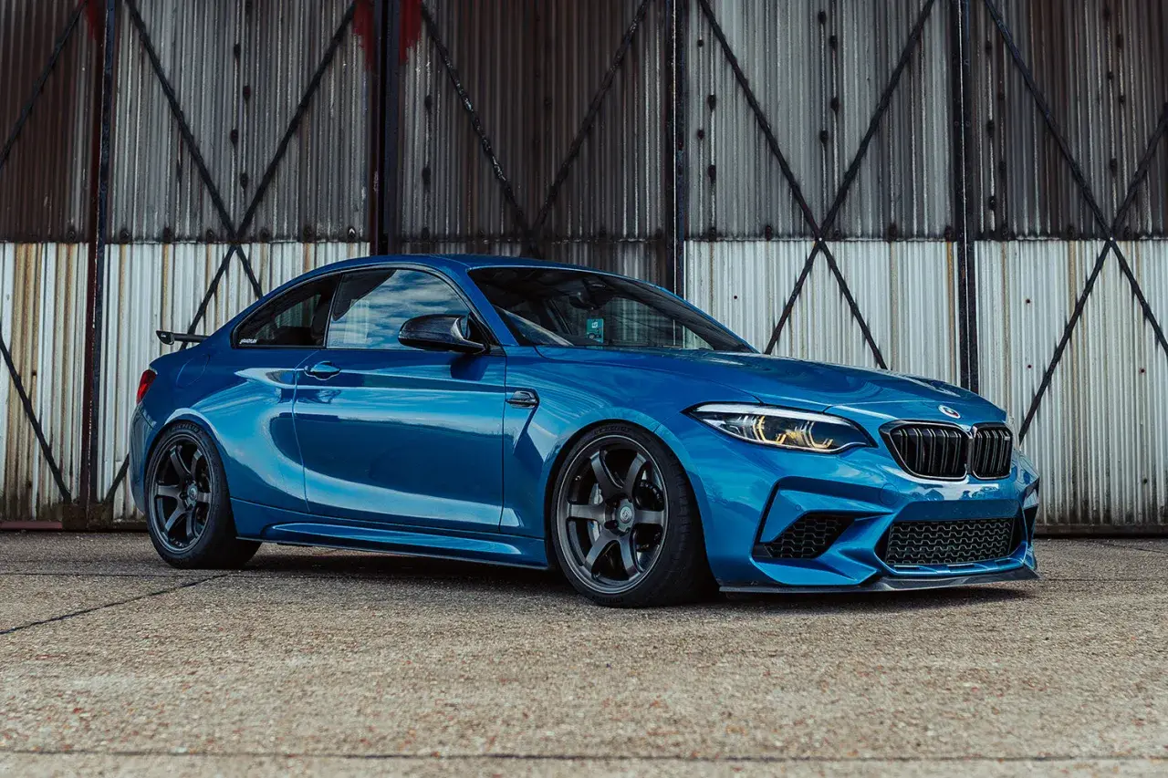 Ein blauer BMW F87 M2 mit dunklen Felgen steht vor einer rostigen Metallwand.