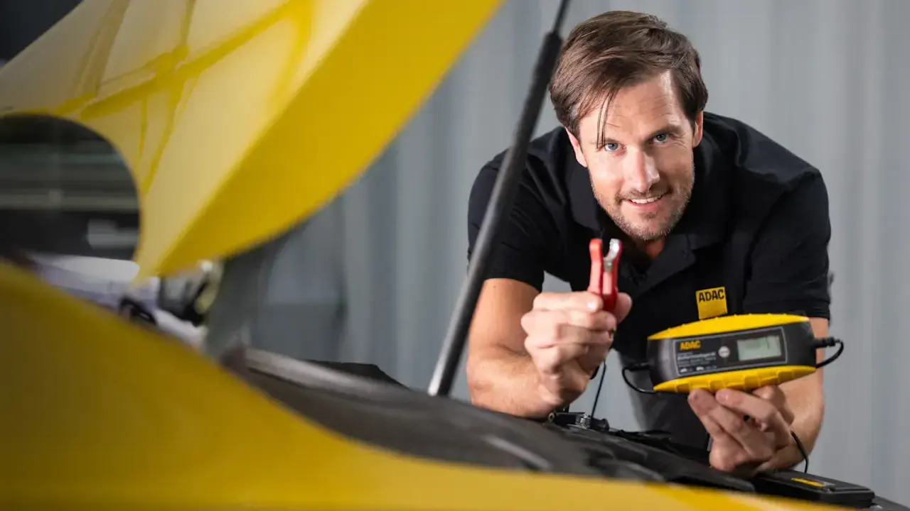 Mann mit ADAC-Kleidung lädt eine Autobatterie im eingebauten Zustand mit einem gelben Ladegerät.