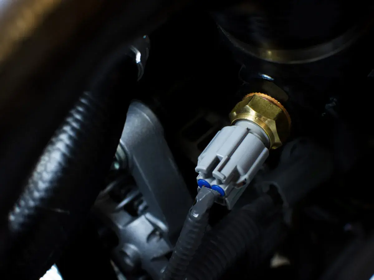 Ein p0118 vw Sensor mit grauem Stecker und Messinggewinde, eingebettet in dunkle Motorraumkabel.