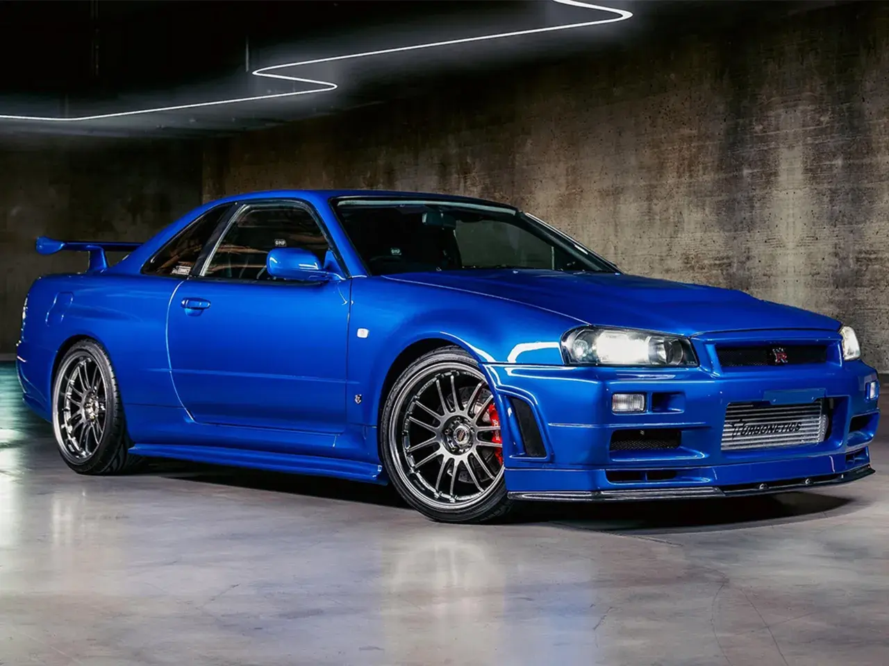Ein blauer Nissan Skyline R34 GT-R mit Spoiler und auffälligen Felgen steht in einer Garage.