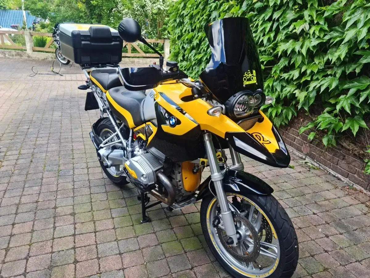 Gelbe BMW 1200 GS mit schwarzer Verkleidung und Gepäckbox, bereit für Abenteuer auf gepflastertem Weg vor grüner Wand.